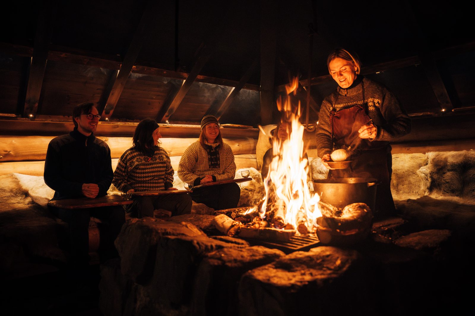 Guests on reindeer hides around the fire in Barta, the cook turning bread by the flames