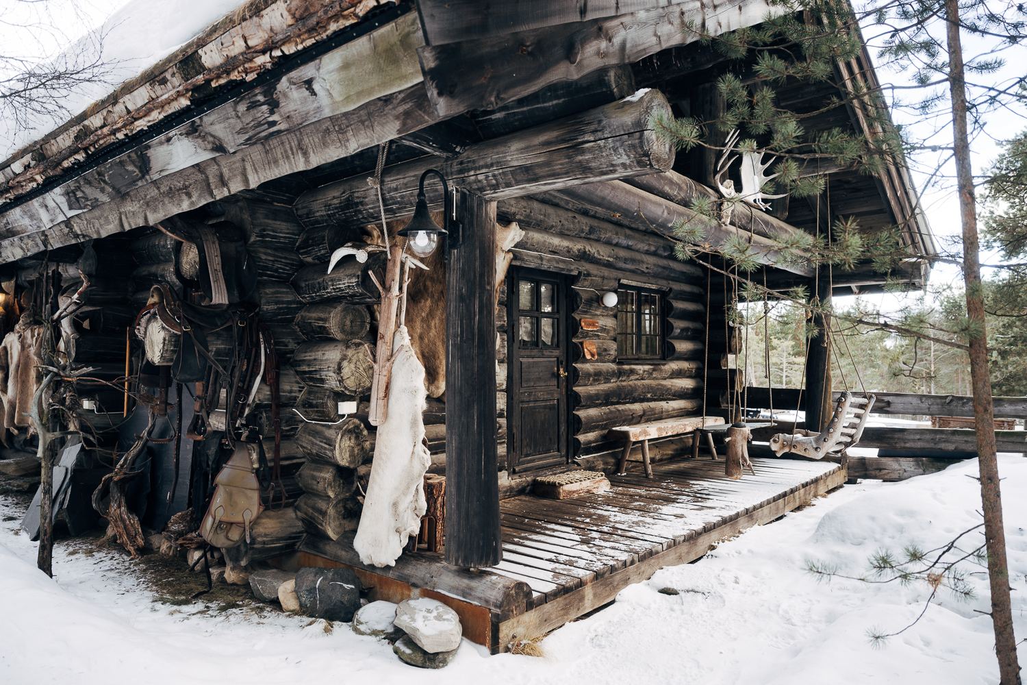 A dark log cabin in deep snow, hides and tools hung along its wall