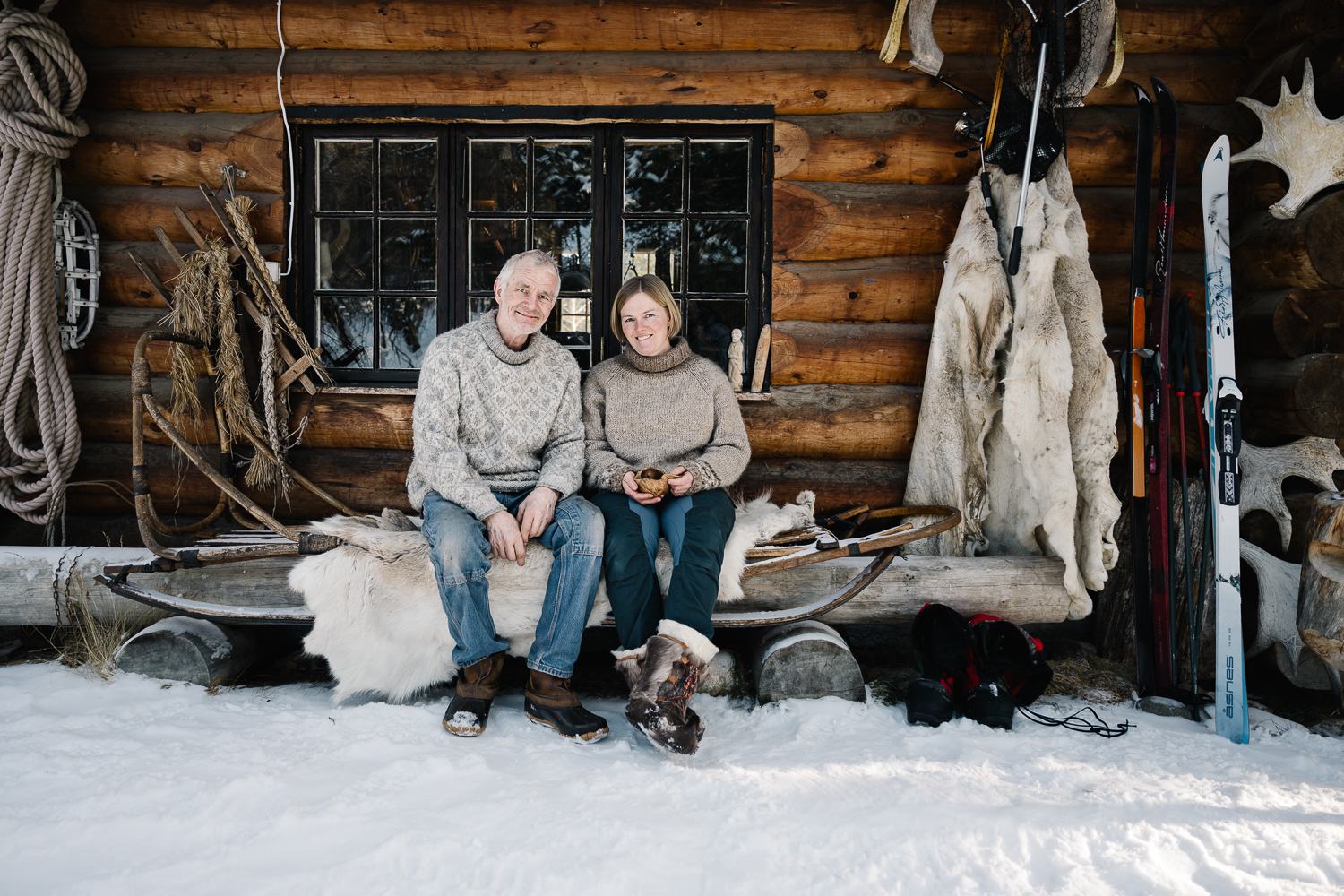 Two people sitting on a sled outside a log cabin in snow