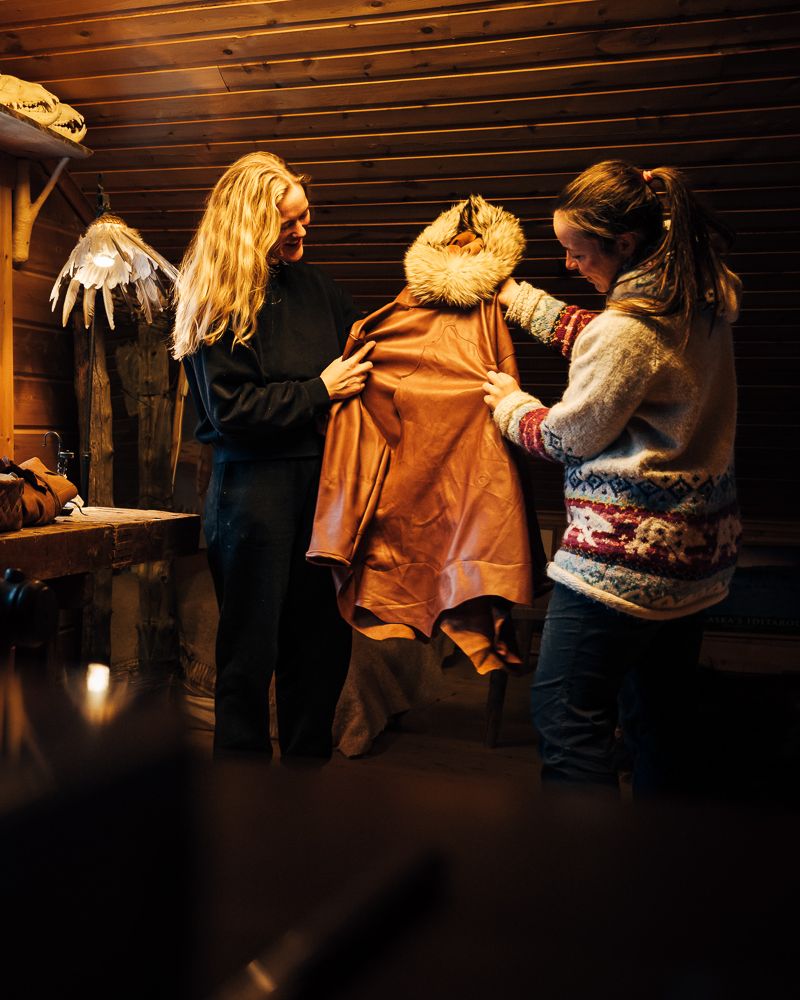 Two women holding up a finished leather anorak with a fur hood in the workshop