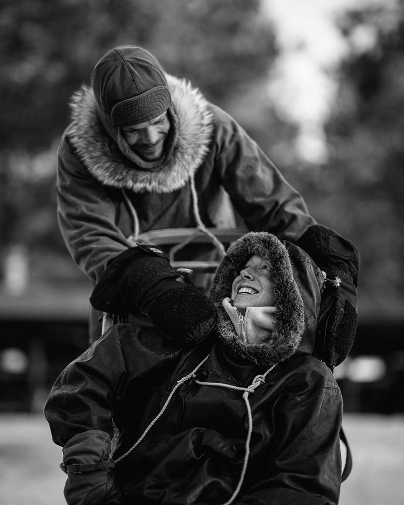 A guide leaning over a laughing passenger tucked into the sled, black and white