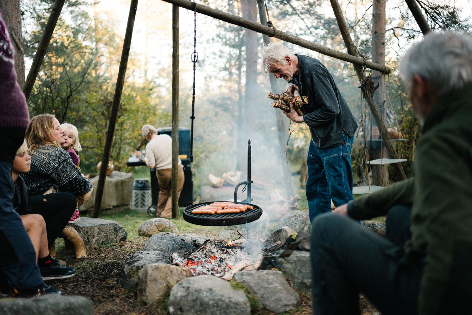 A pan lifted over the fire pit under the tripod, guests on the stones around it