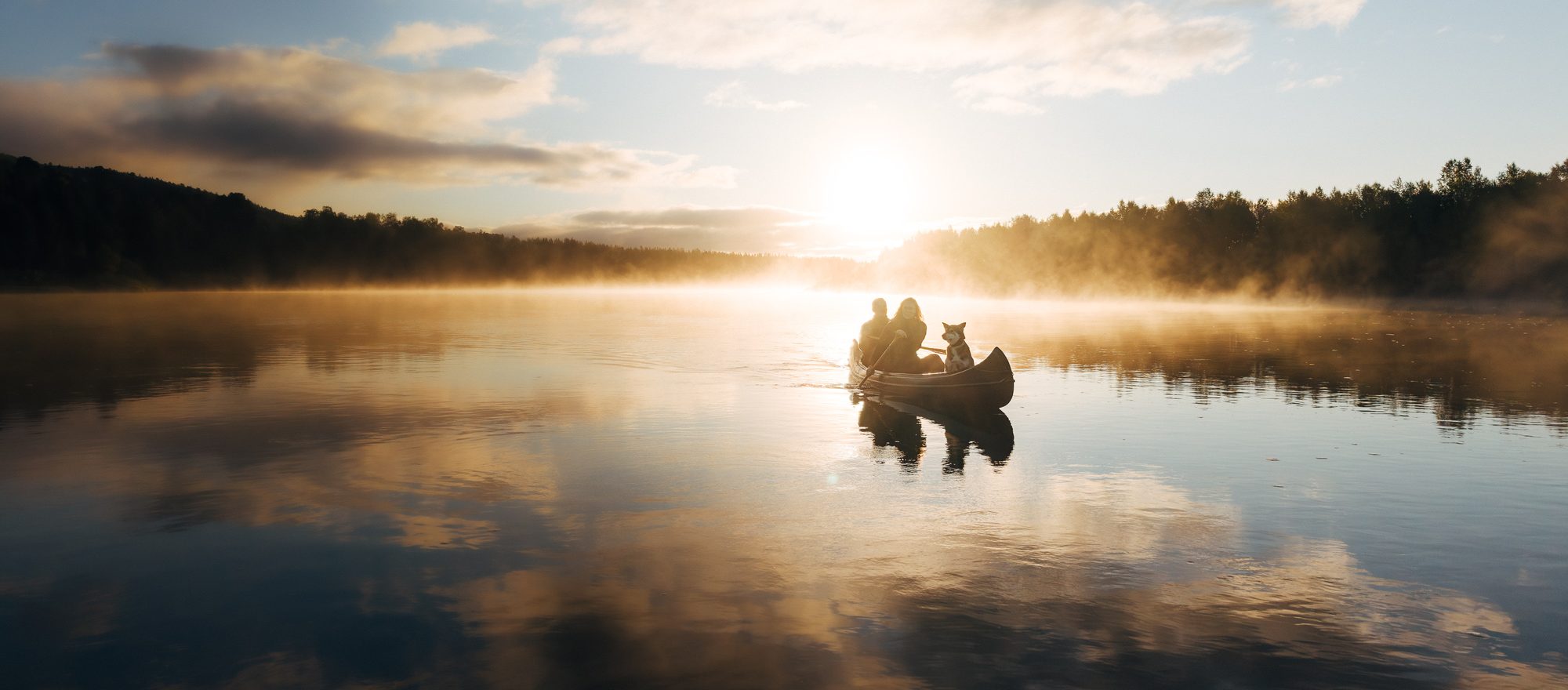 Two people and a dog in a canoe on the Kárášjohka at sunrise, mist lying over the water and the forest dark along both banks