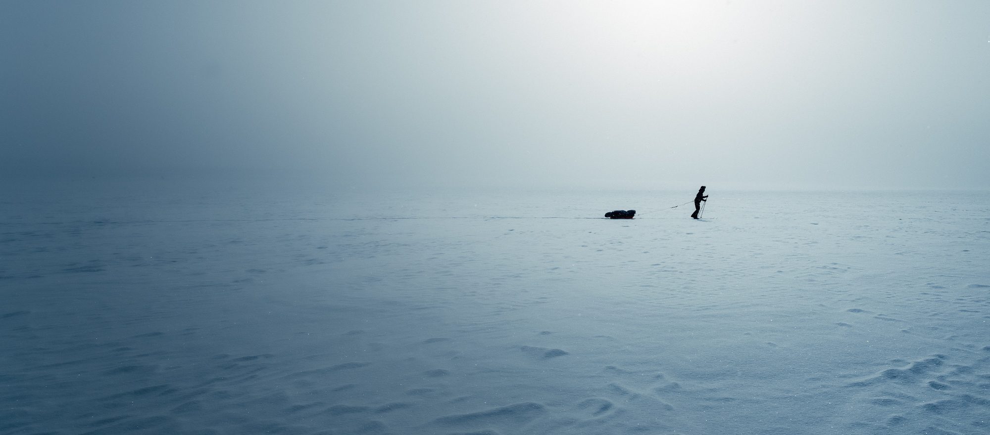 A single skier pulling a pulk across an empty blue-grey plateau, the horizon lost in mist