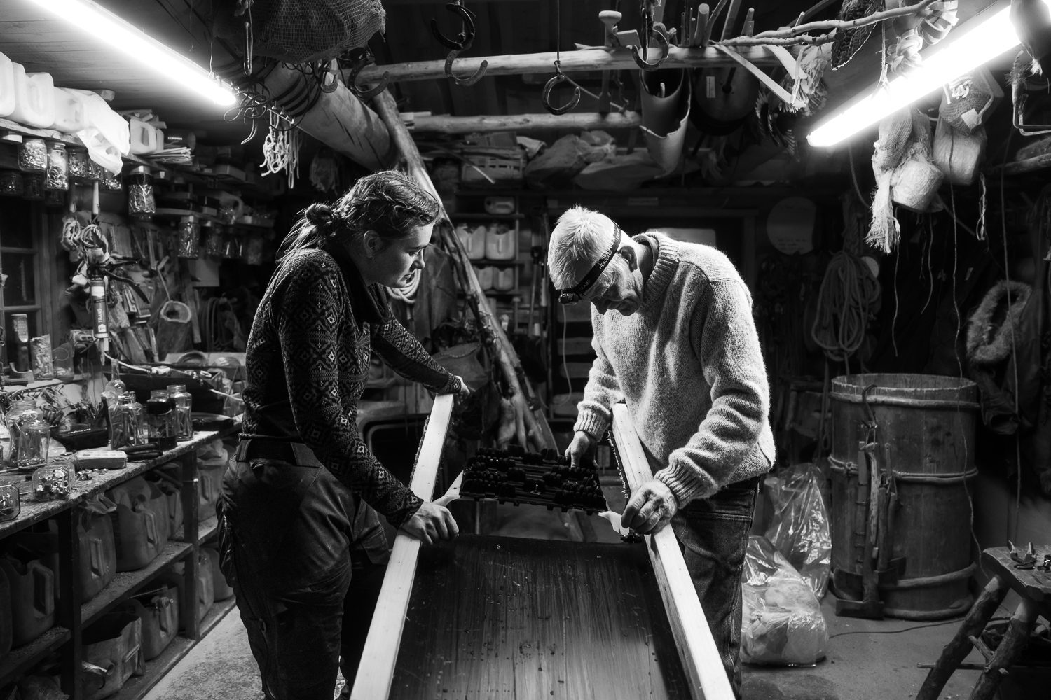 Two people working a long plank of wood in the lodge workshop, tools and rope hanging from the ceiling