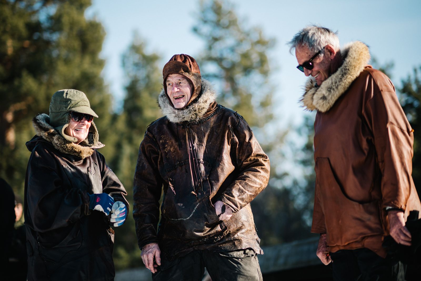 Three people in fur-hooded leather anoraks laughing together among the pines in the low winter sun
